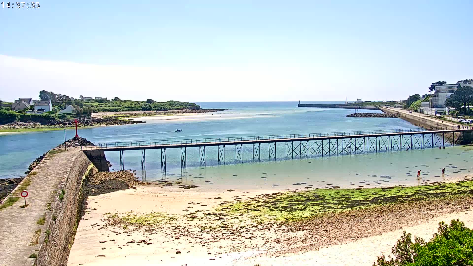 A long pier extends over shallow, clear water toward a distant shoreline under a sunny sky, with a sandy beach and rocky areas visible in the foreground.