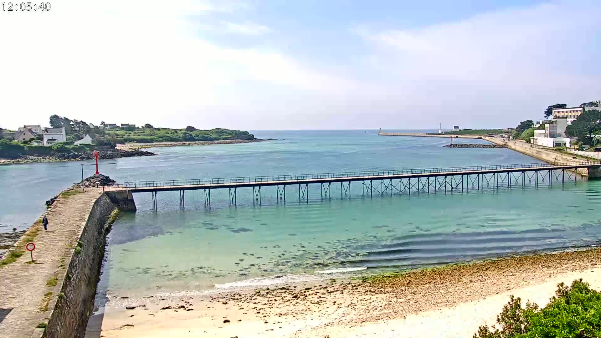 A long pier stretches across calm, clear turquoise water toward a small island and a sandy beach under a mostly sunny sky.