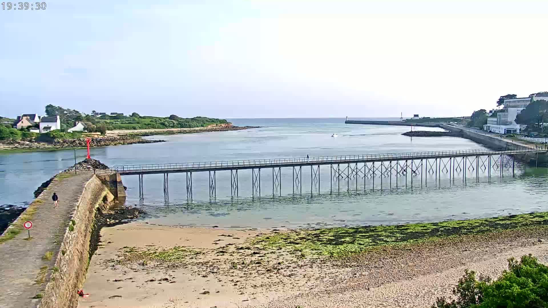 A long pier extends over calm, shallow water towards a small town on a sunny day, with a sandy beach and green vegetation visible along the shore.