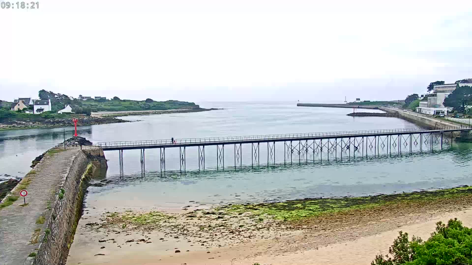 A long pier extends over calm, shallow water toward a small town under an overcast sky, with a sandy beach visible in the foreground.