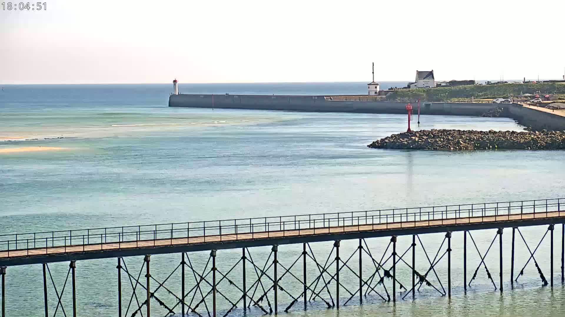 A long pier extends over calm, light-blue water toward a harbor wall with a lighthouse and small building, under a clear sky.