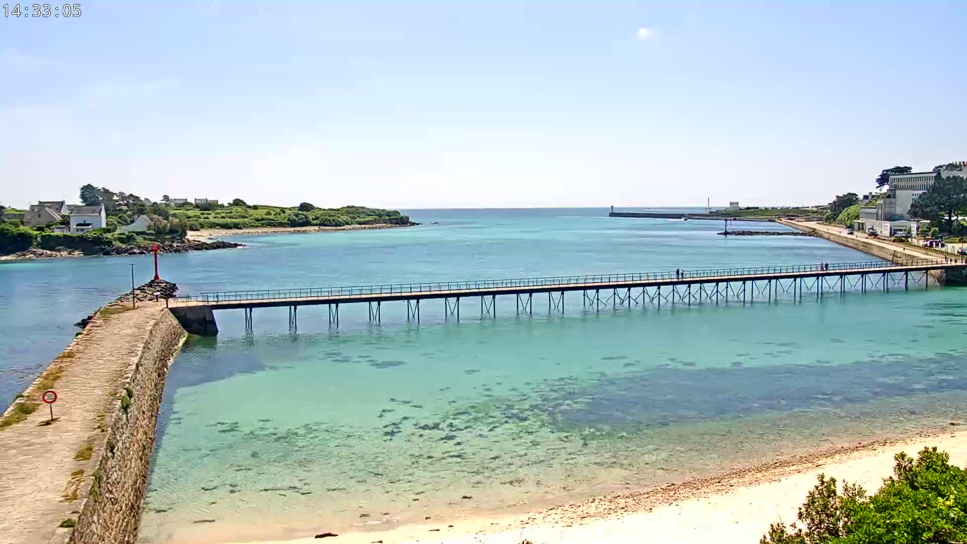 A long pier extends over clear, shallow turquoise water toward a distant shore on a sunny day.