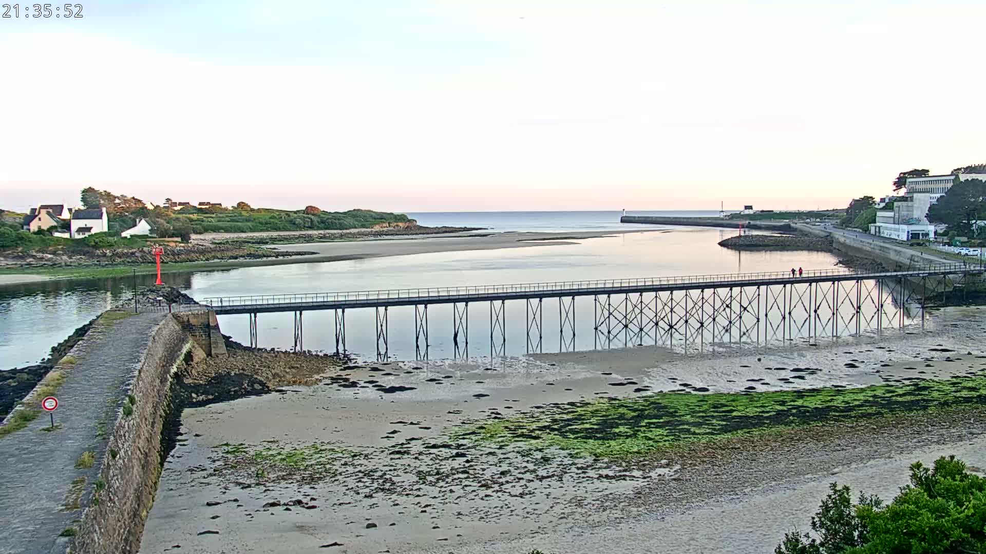 A long pier stretches across a calm bay at low tide, under a pale evening sky, with buildings and green vegetation visible on both sides of the water.
