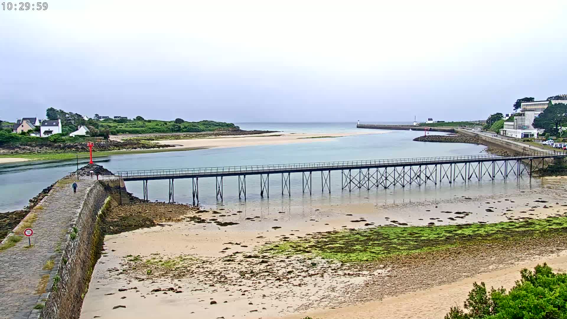 A long pier stretches across a calm, shallow waterway between a sandy beach and a shoreline with buildings under an overcast sky.