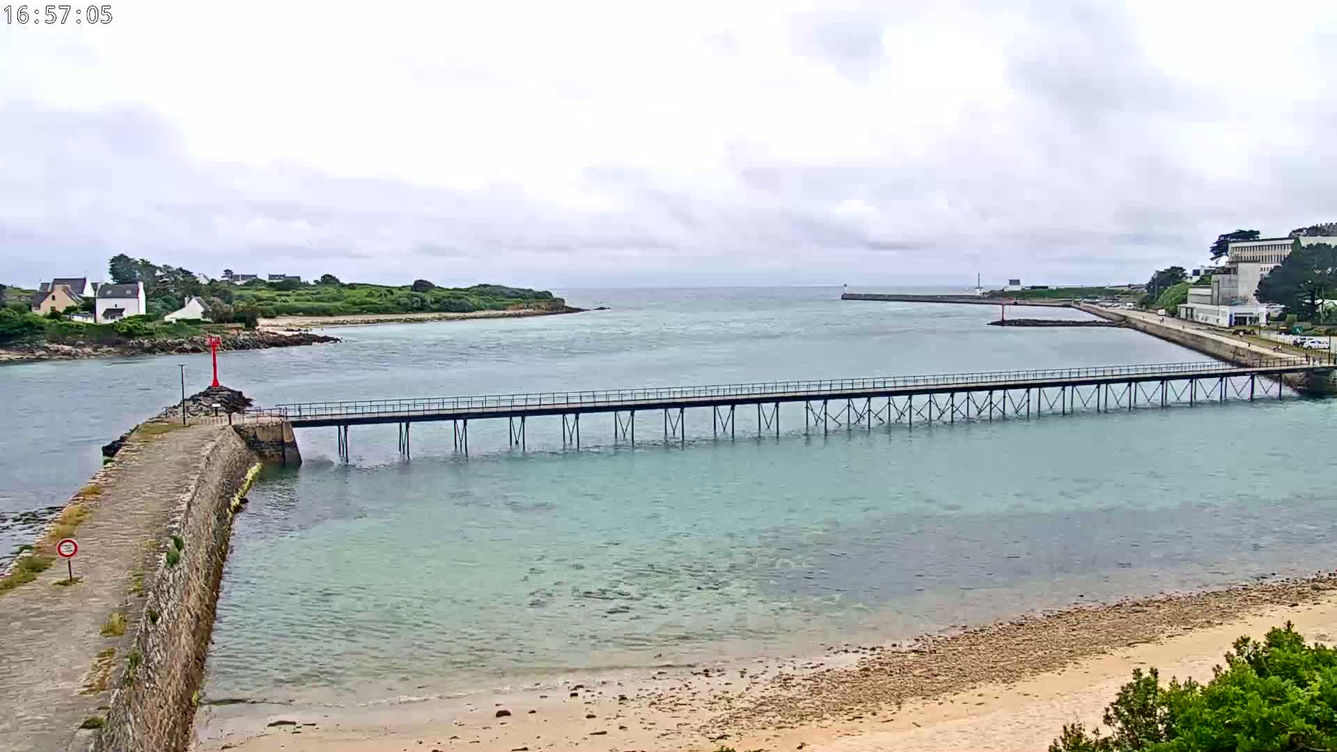 A long pier stretches across calm, light-blue water under an overcast sky, connecting a stone jetty to land with buildings and green vegetation in the distance.