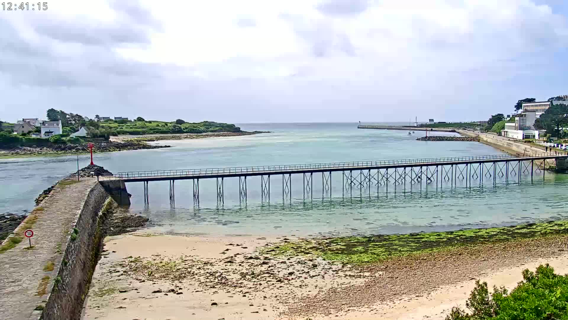 A long pier extends over shallow, clear water toward a small island under a partly cloudy sky.
