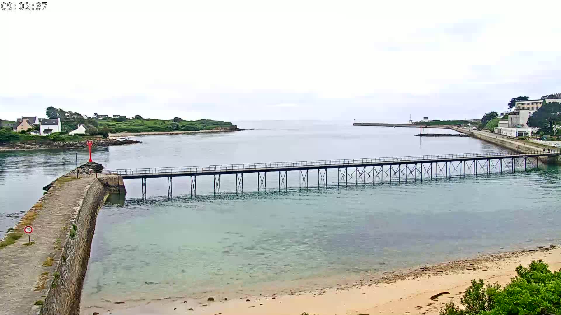 A long pier stretches across calm, light-green water toward a small town and island under an overcast sky.