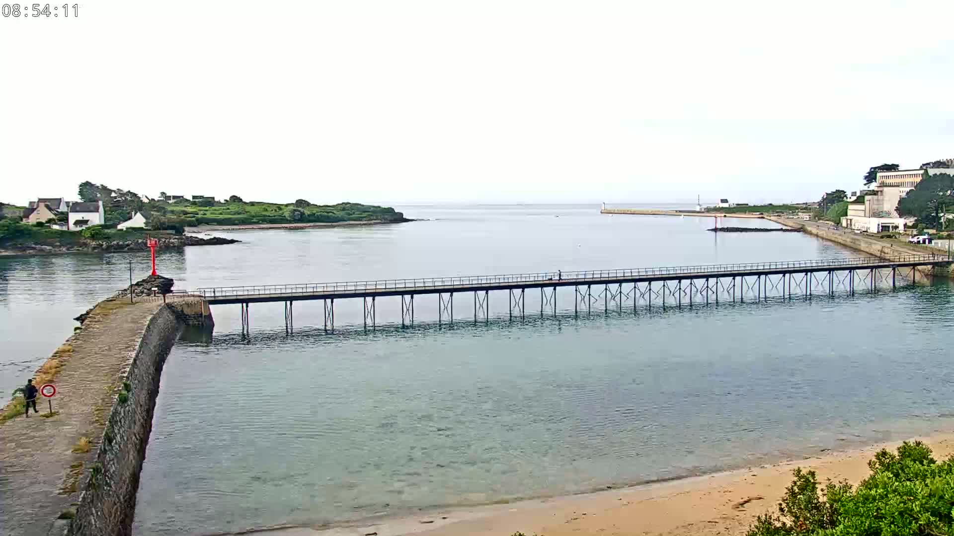 A long pier stretches across calm, light-blue water between two shorelines, one featuring a small village and the other a building, under an overcast sky.