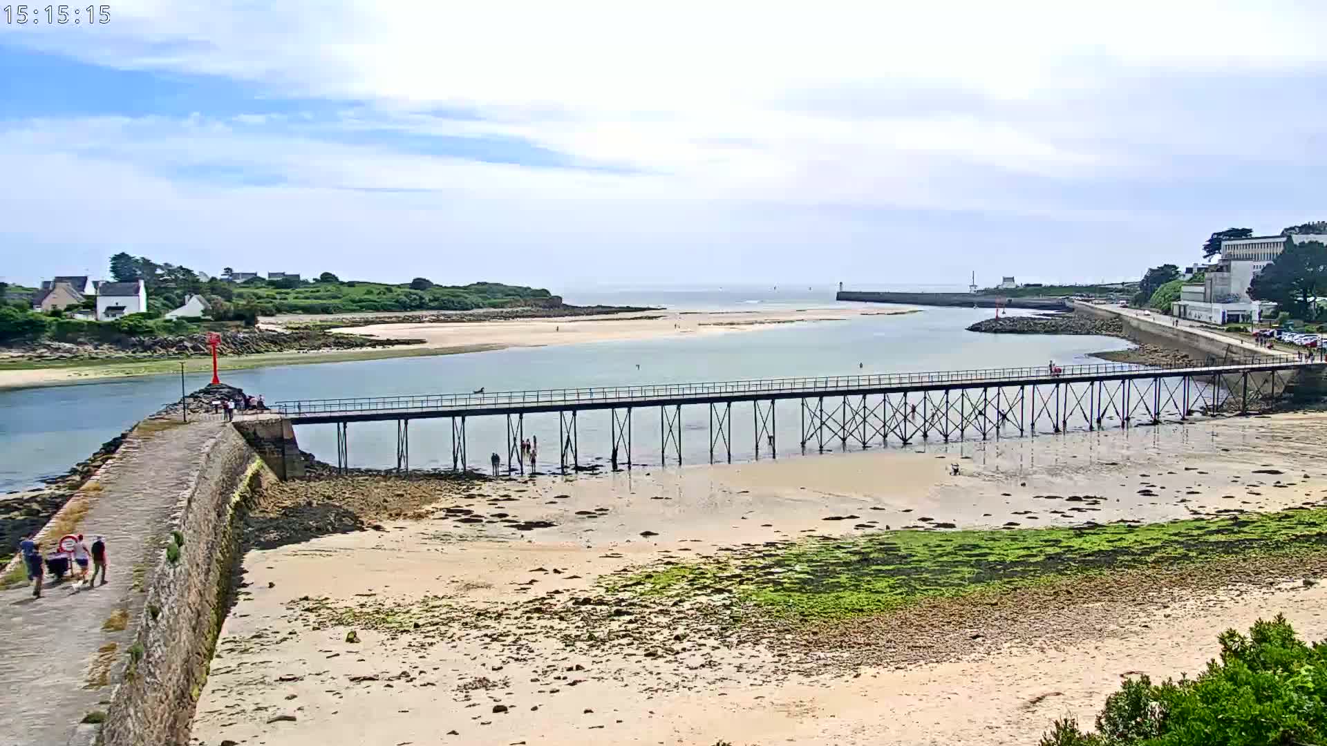 A long pier extends over a calm, shallow body of water, connecting a stone wall to the opposite shore under a partly cloudy sky.