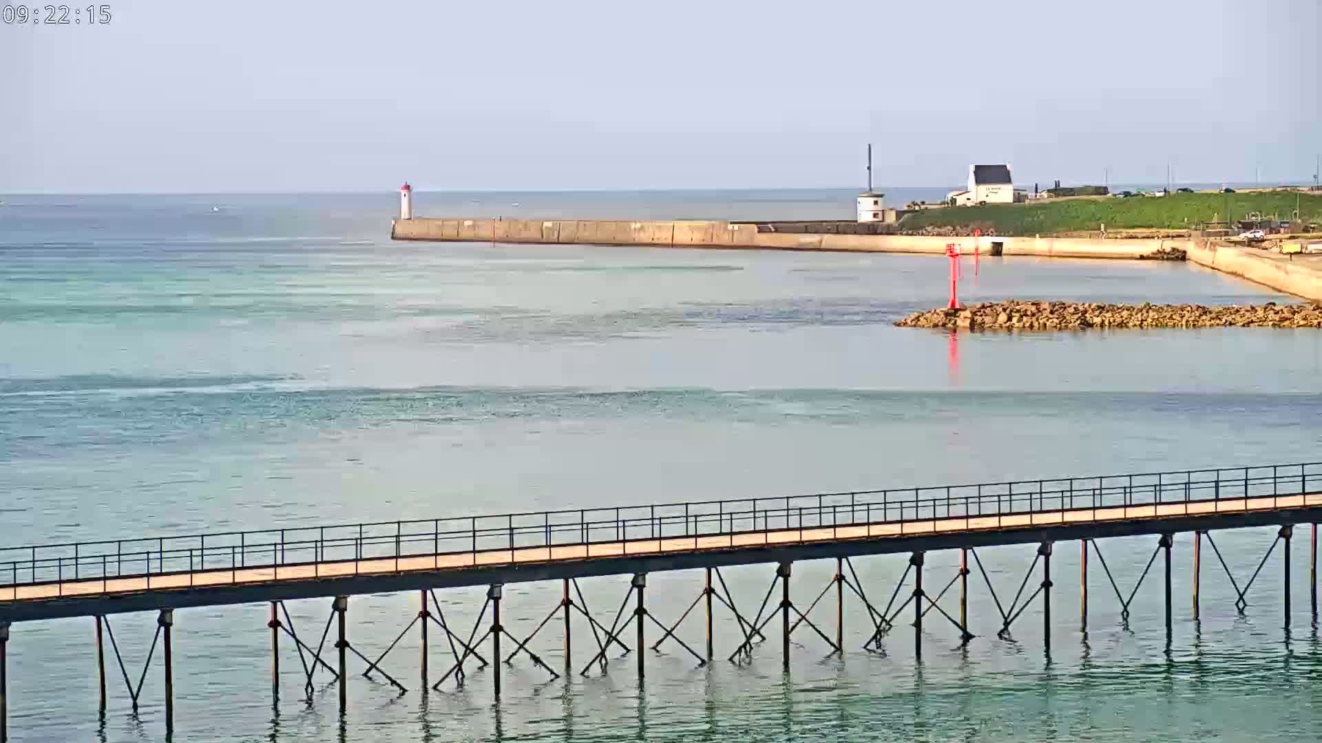 A long pier extends over calm, light-blue water toward a harbor with a lighthouse and a small building on a sunny day.