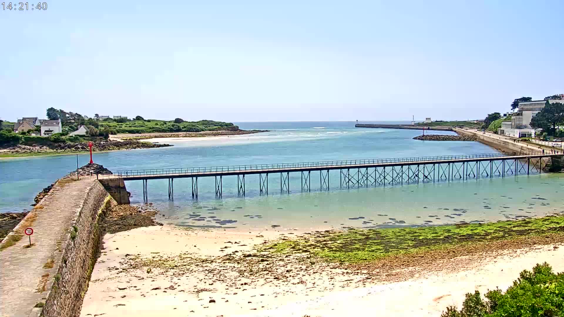 A long pier stretches across calm, shallow water toward a small town on a sunny day, with sandy shores and green seaweed visible in the foreground.