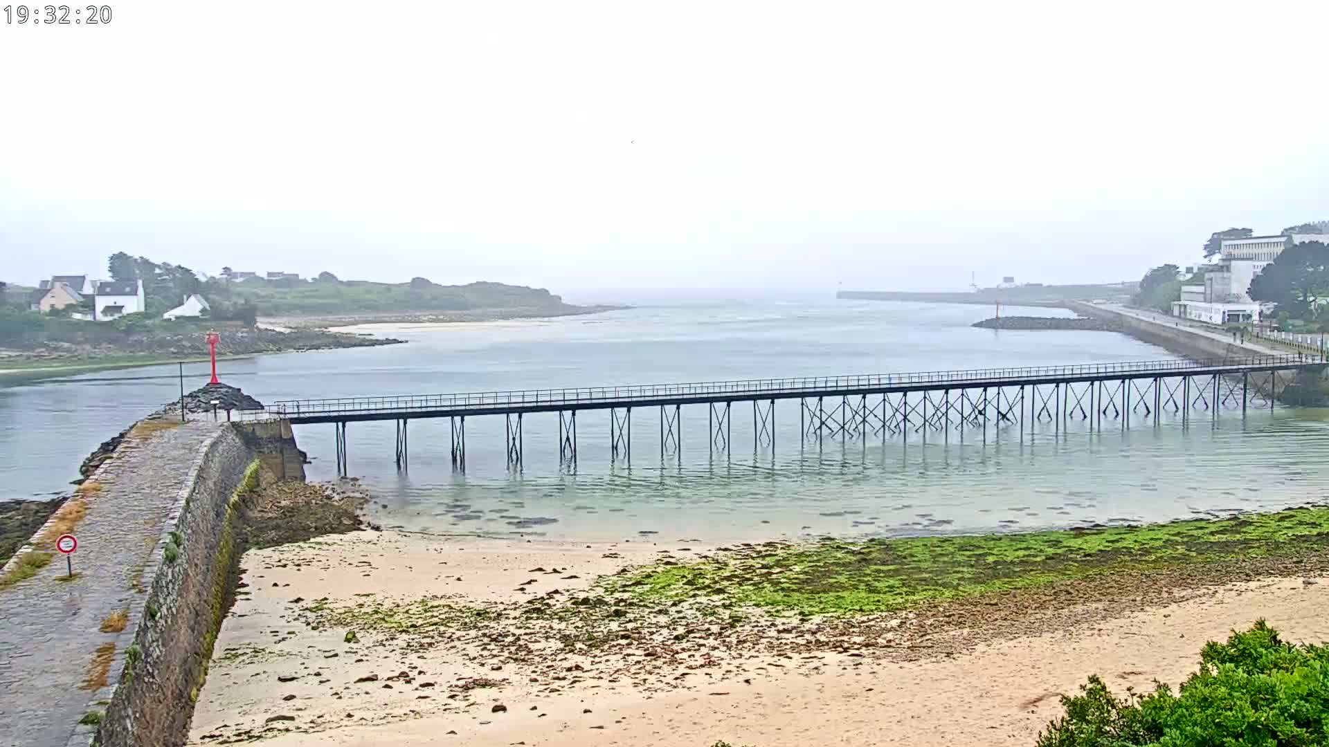 A long pier stretches across calm, shallow water between two landmasses on an overcast day, with a sandy beach and green algae visible at low tide.