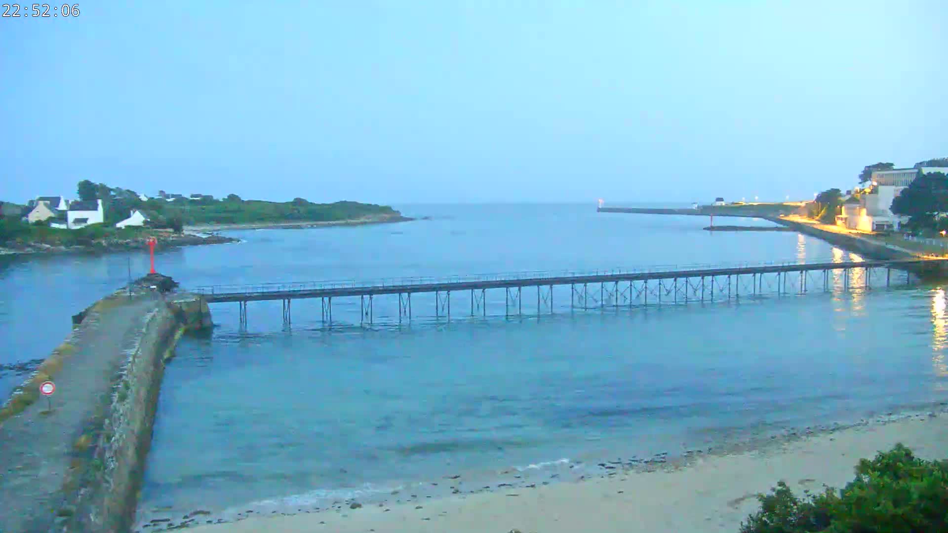 A long pier extends across calm, blue water toward a landmass at dusk, with a sandy beach and buildings visible on the shore.
