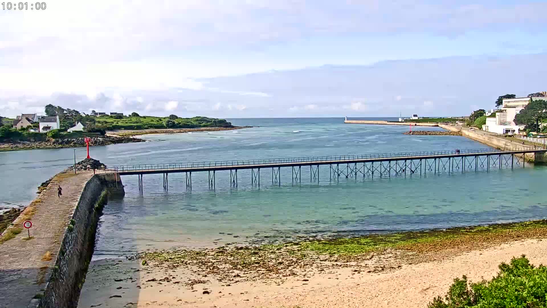 A long pier stretches across calm, clear water toward a small island under a partly cloudy sky.