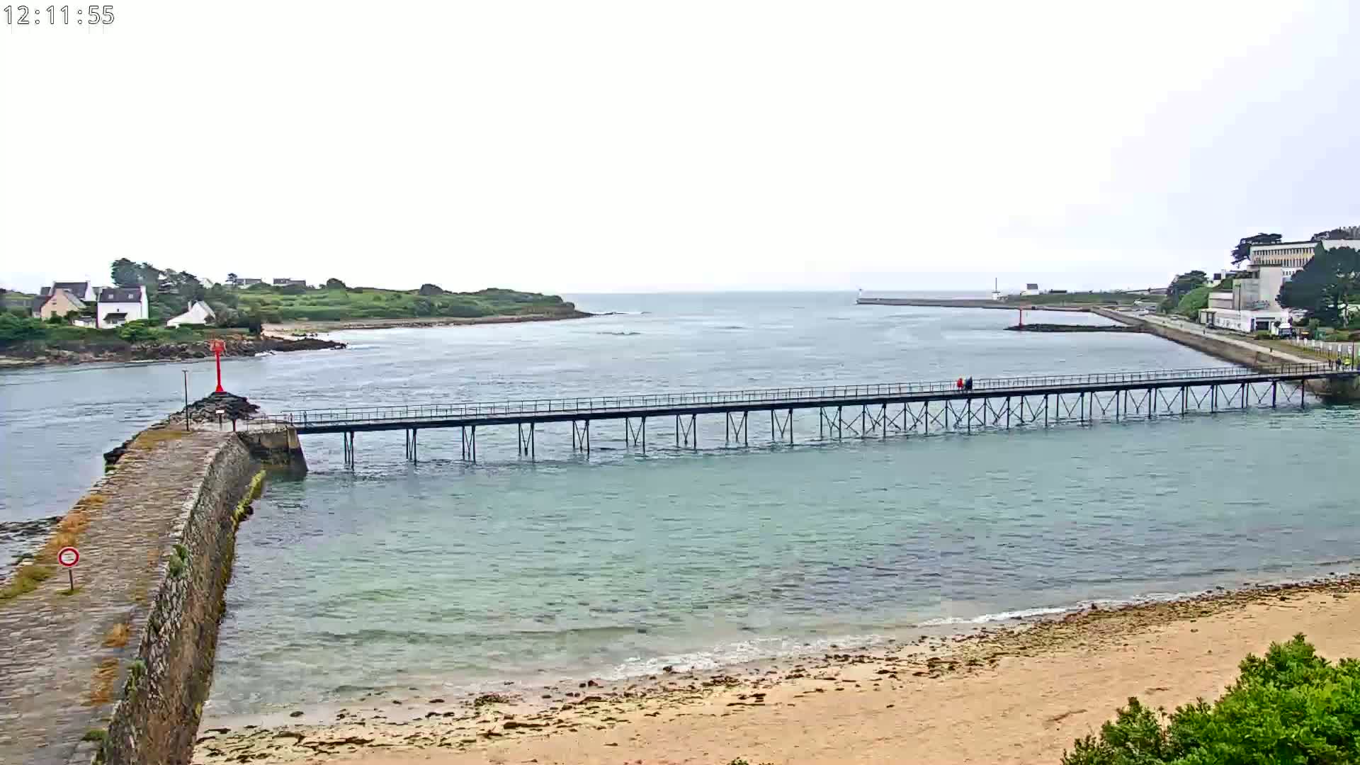 A long pier stretches across calm, light-blue water under an overcast sky, connecting a stone jetty to a distant shore with buildings.
