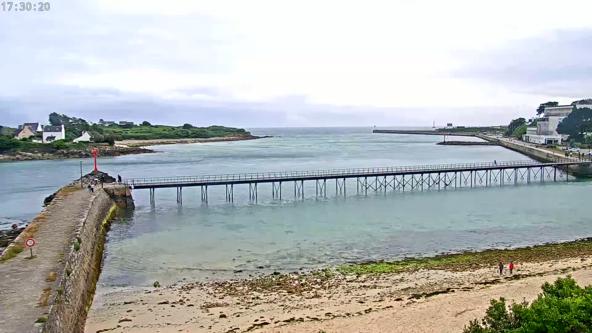 A long pier extends over calm, light-blue water toward a distant shoreline under an overcast sky.