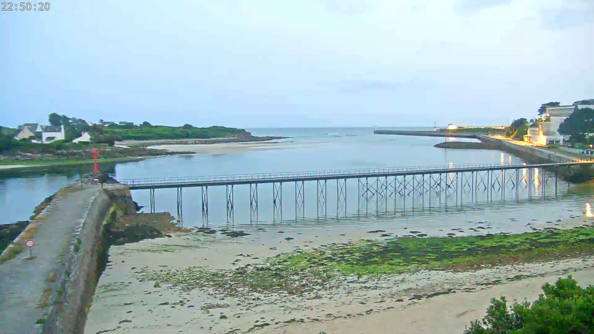 A long pier stretches across calm water separating two stretches of land, one with buildings, under a pale, overcast sky.