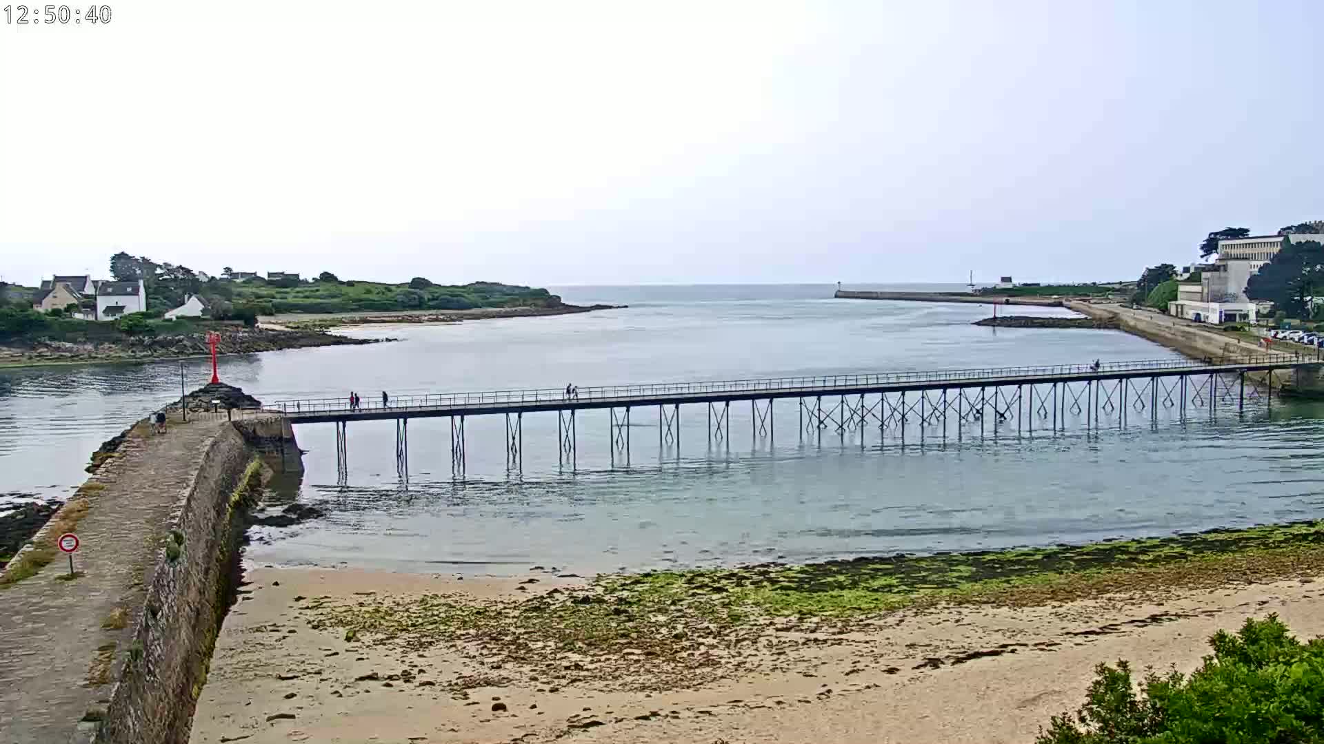 A long pier stretches across calm water separating a sandy beach and a small town from a larger landmass under an overcast sky.