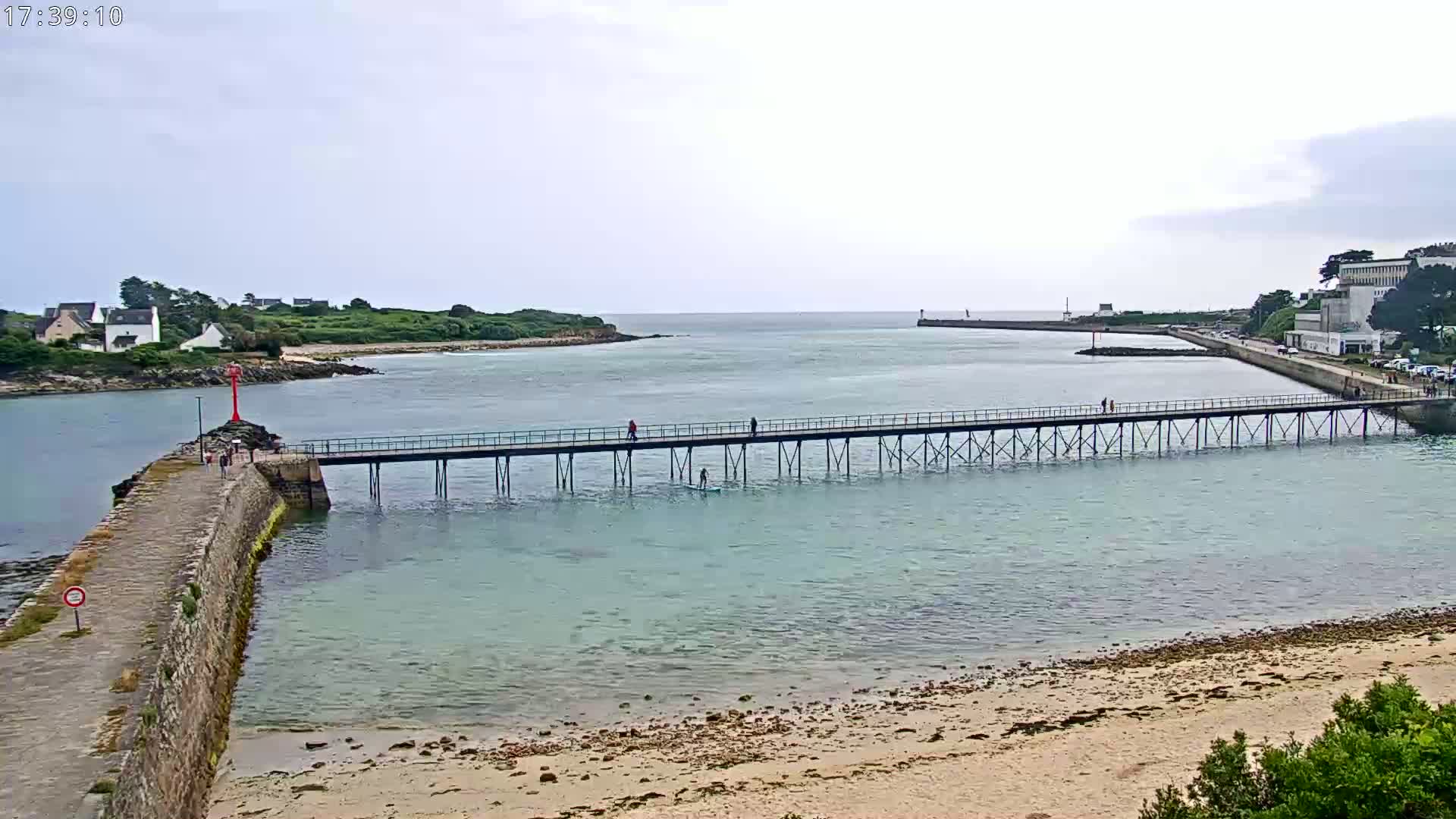 A long pier extends over calm, light-blue water toward a shoreline with buildings and a small island under an overcast sky.