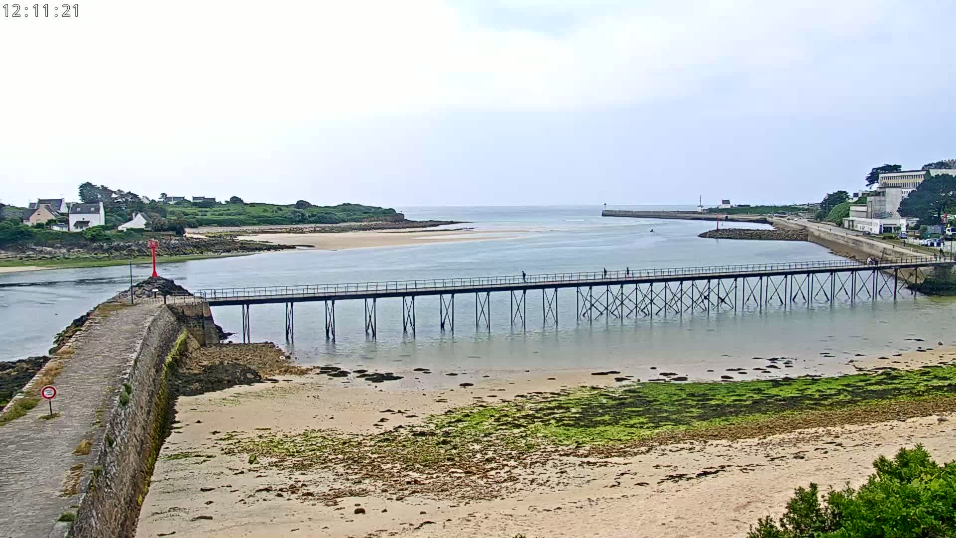 A long pier extends over shallow, calm water toward a small town on a partly cloudy day, with sandy beaches and green seaweed visible along the shoreline.