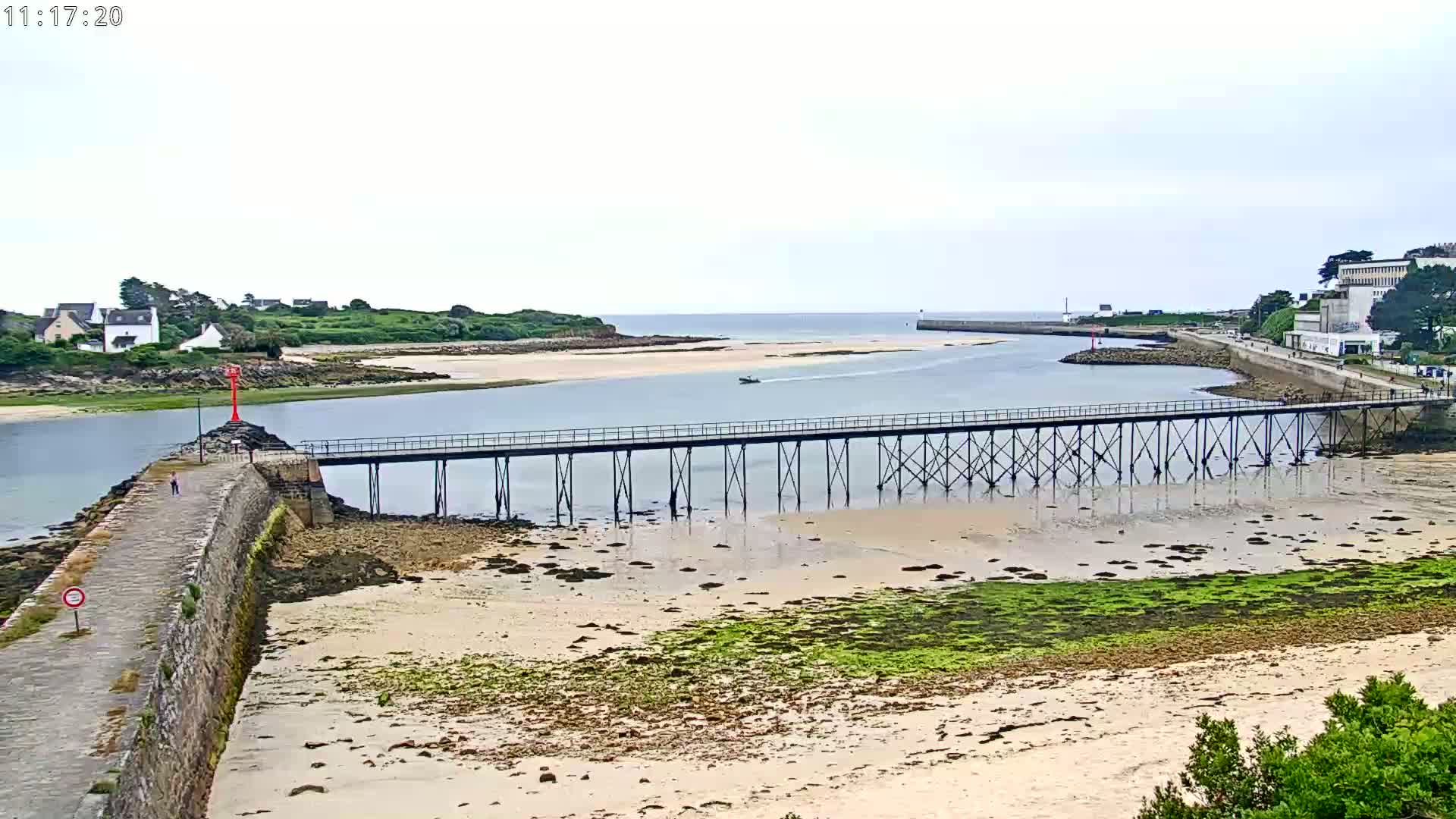 A long pier extends over a shallow, sandy waterway under an overcast sky, with a small boat visible in the distance.