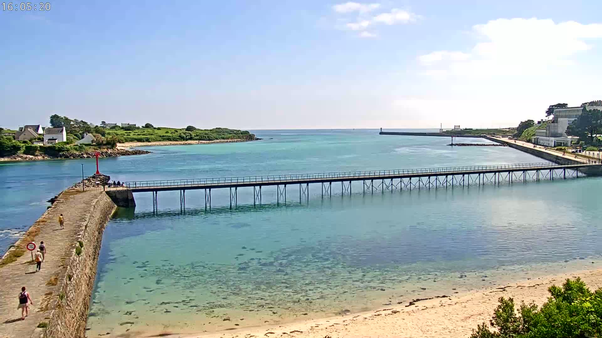 A long pier stretches across clear, shallow turquoise water toward a distant shoreline under a mostly sunny sky, with people walking along the pier and a stone walkway leading to it.