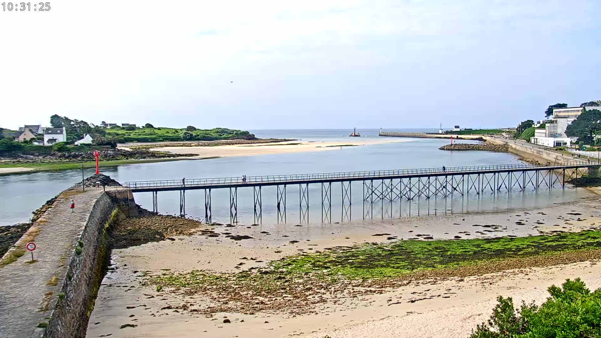 A long pier extends over calm water toward a sandy beach and small island, under a partly cloudy sky.