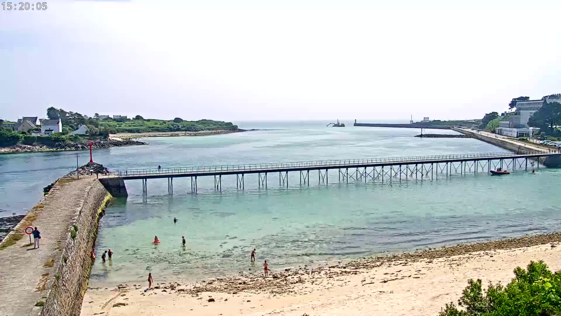 A long pier stretches across calm, shallow turquoise water toward a small town and distant land, with people swimming and sunbathing on a sandy beach under a bright, mostly clear sky.