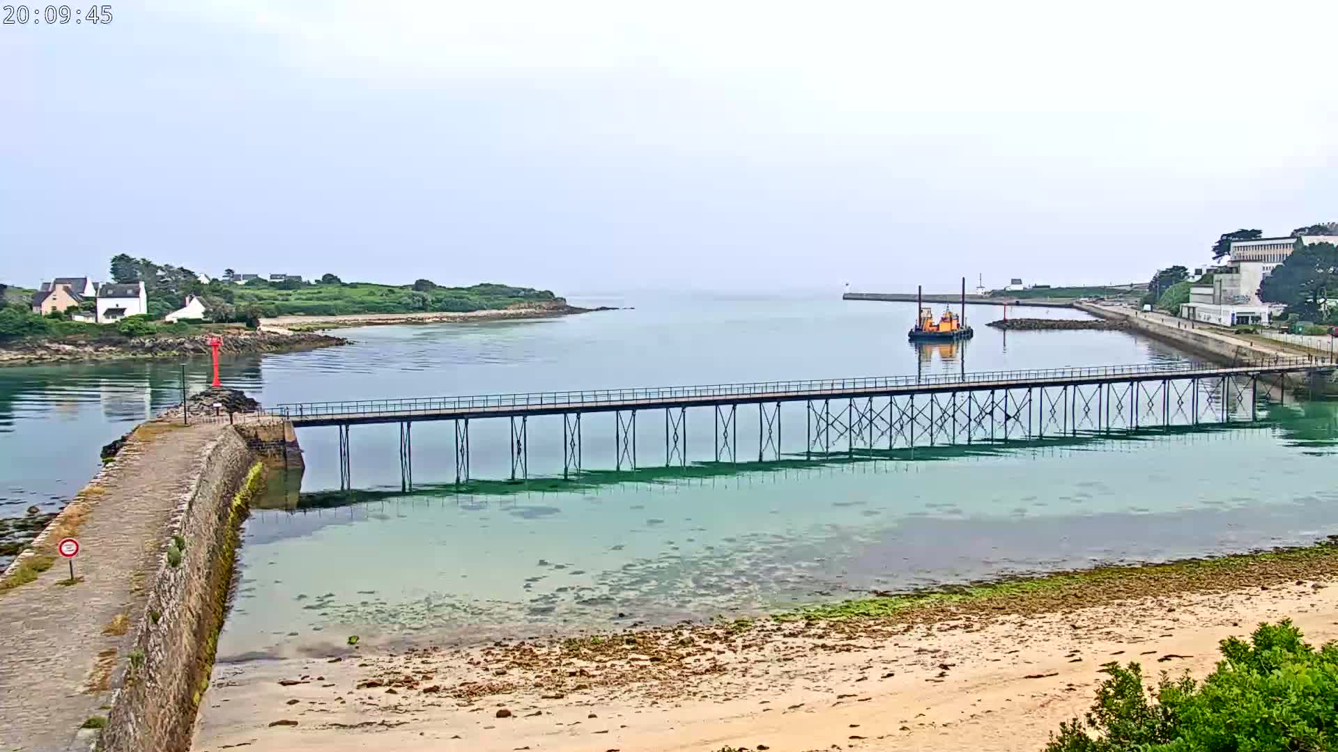 A long pier stretches across calm, shallow, green-blue water toward a small boat, with a hazy sky and a sandy beach in the foreground.