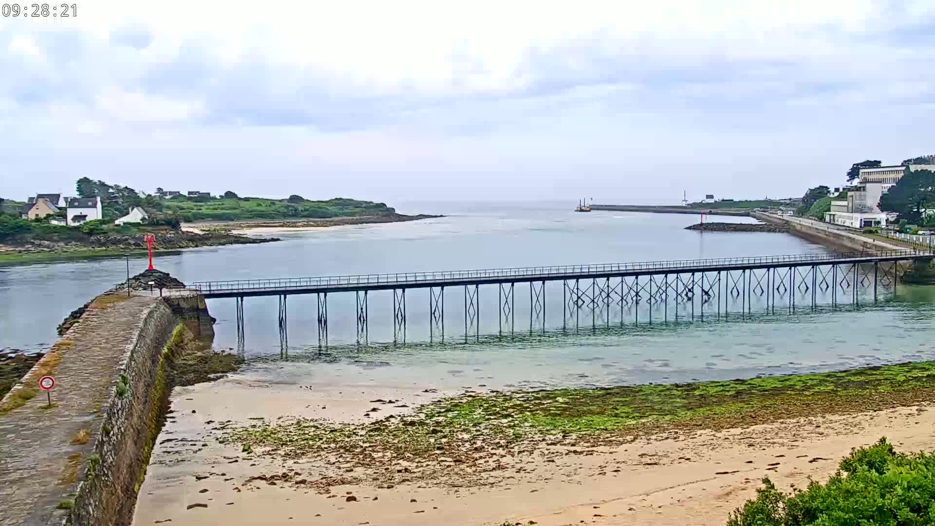 A long pier stretches across calm, shallow water, connecting a stone wall to a landmass with buildings, under a mostly cloudy sky.