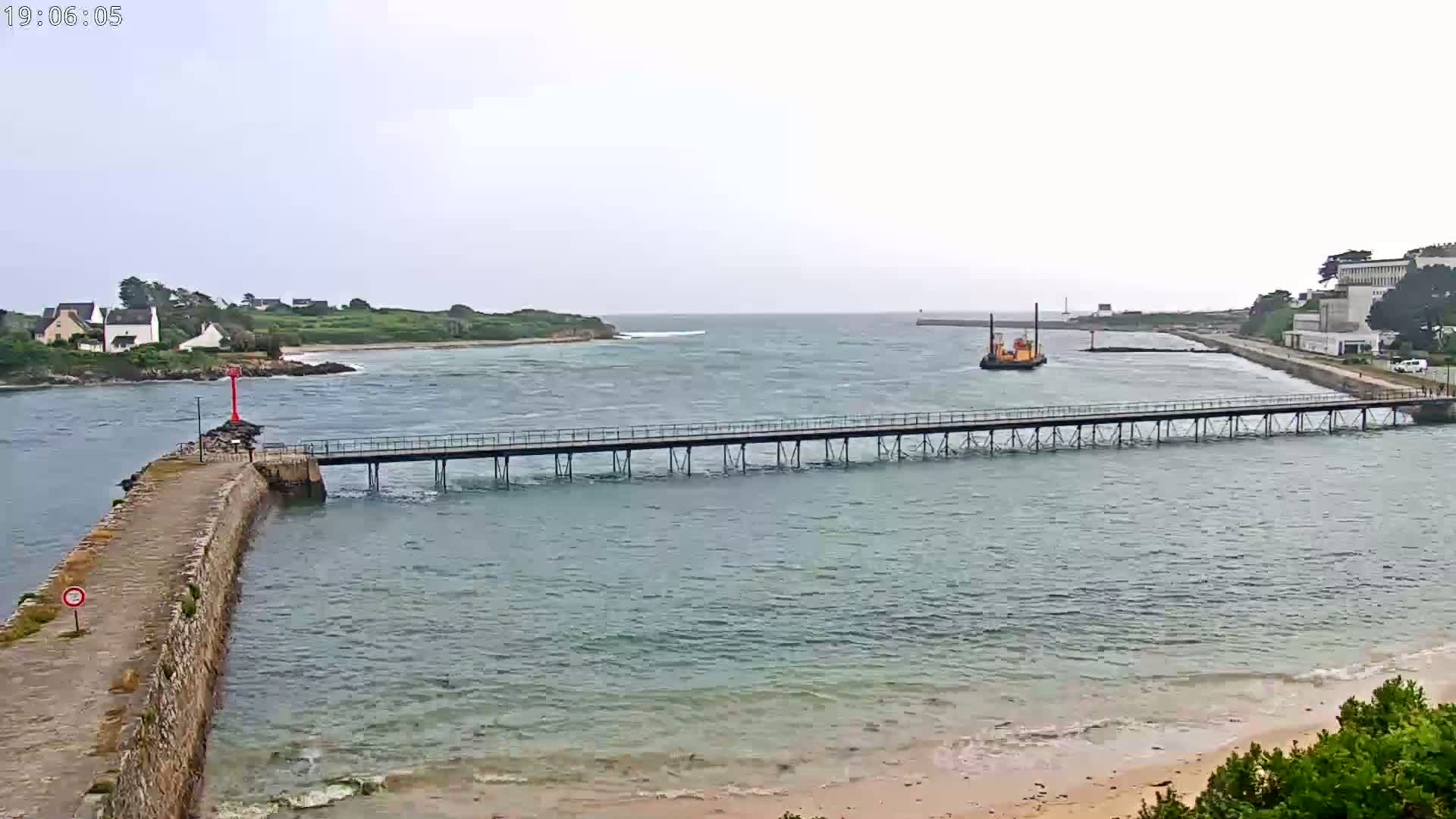 A long pier stretches across a calm body of water under an overcast sky, connecting a stone jetty to a distant shore with buildings and a small boat.