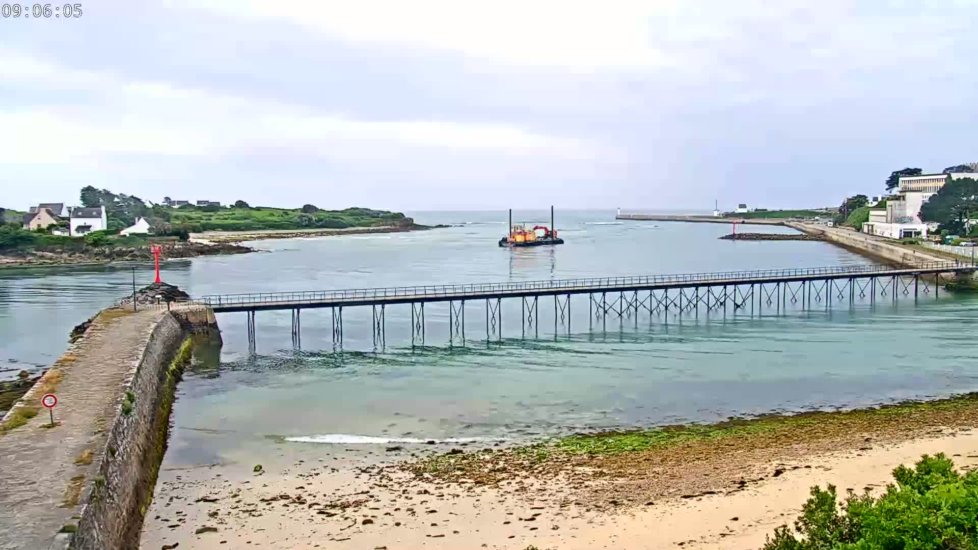 A long pier extends over calm, shallow water toward a small island and a dredging vessel under an overcast sky.