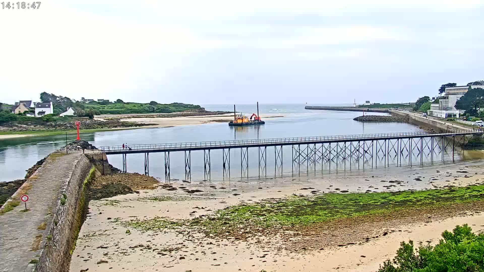 A long pier stretches across calm water toward a small dredging vessel near a shoreline with low-lying buildings and overcast skies.