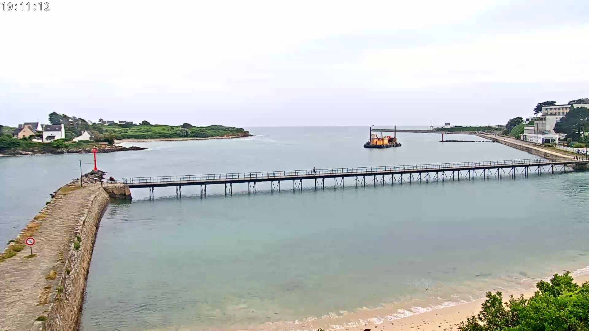 A long pier extends across calm, light-green water toward a small, orange barge, with overcast skies above a coastal scene of buildings and green land.