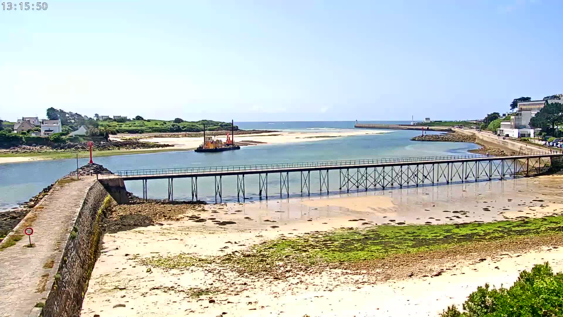 A long pier extends over shallow, sandy water toward a calm inlet, with a small vessel visible in the distance under sunny skies.