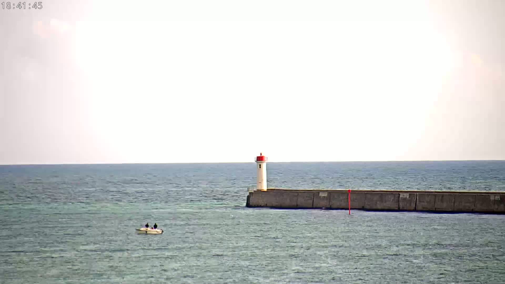 A small boat is on a calm sea near a lighthouse on a breakwater under a mostly clear sky.