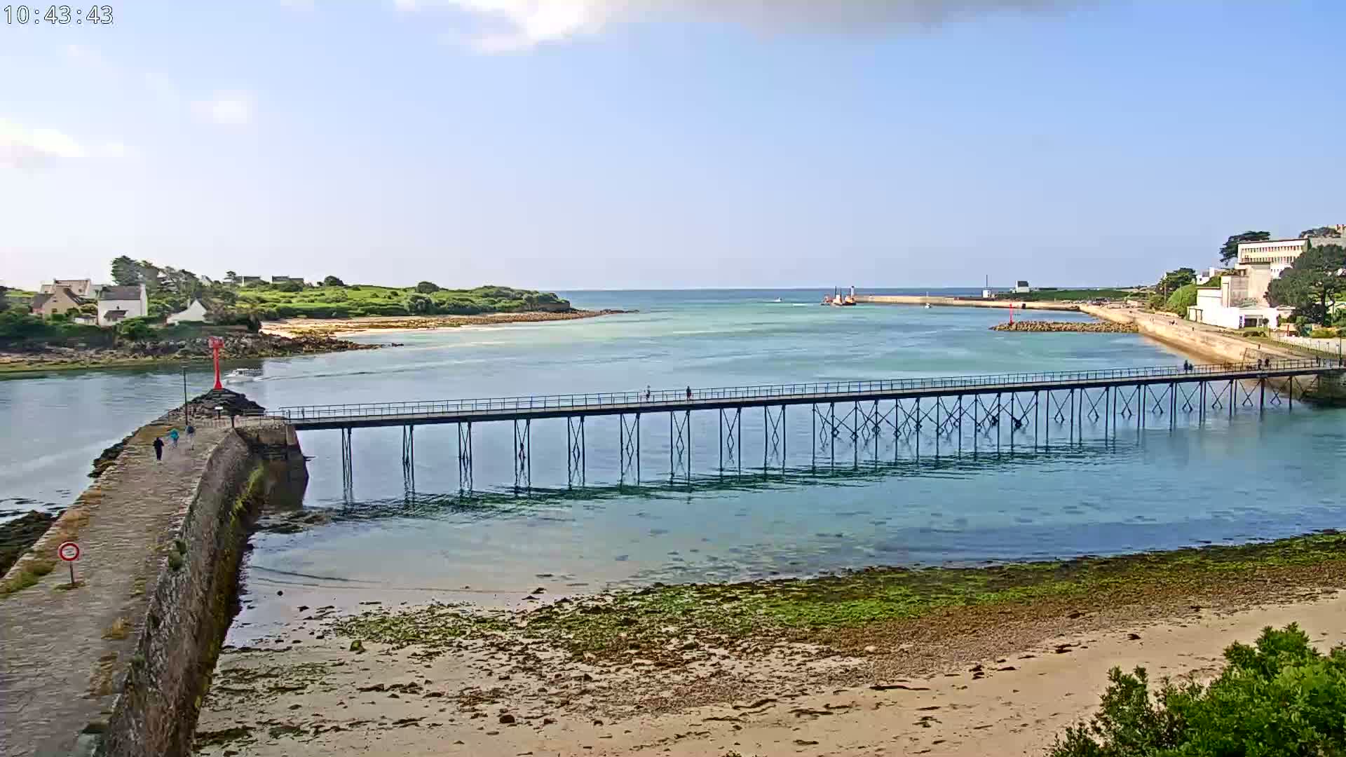 A long pier extends over calm, shallow teal water toward a small island under a mostly sunny sky.