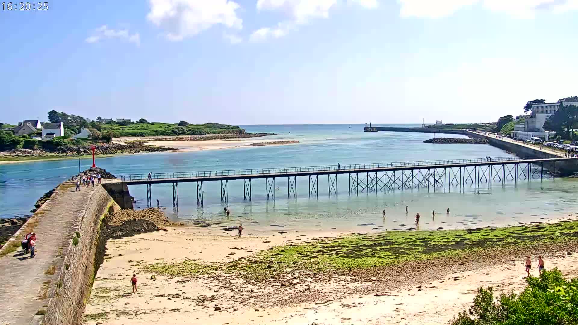 A long pier stretches across shallow, clear water toward a small island on a sunny day, with people walking on the pier and swimming in the water.