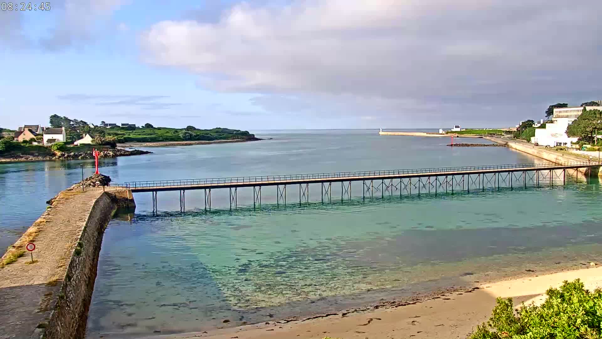 A long pier stretches across shallow, clear water toward a small island on a sunny day, with people walking on the pier and swimming in the water.