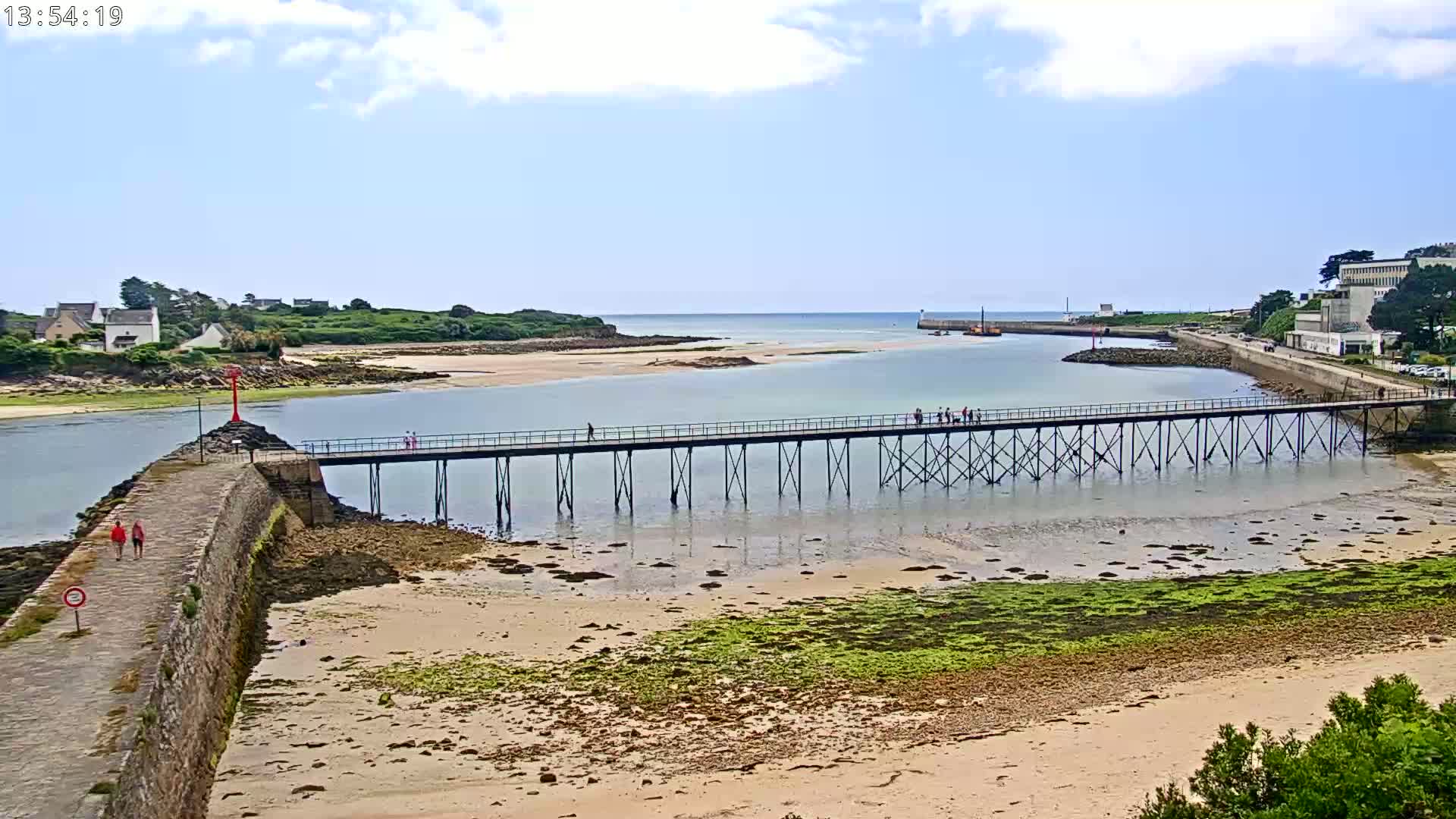 A long pier extends over shallow, sandy water, connecting two landmasses under a partly cloudy sky.