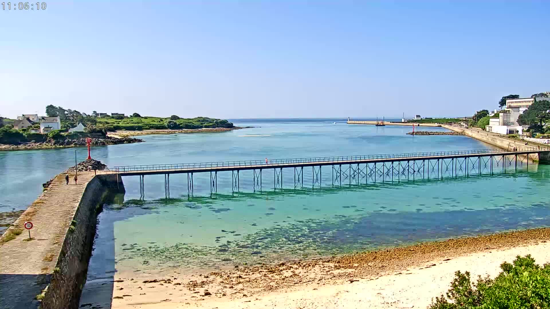 A long pier extends over clear, shallow turquoise water toward a small, tranquil bay on a sunny day.