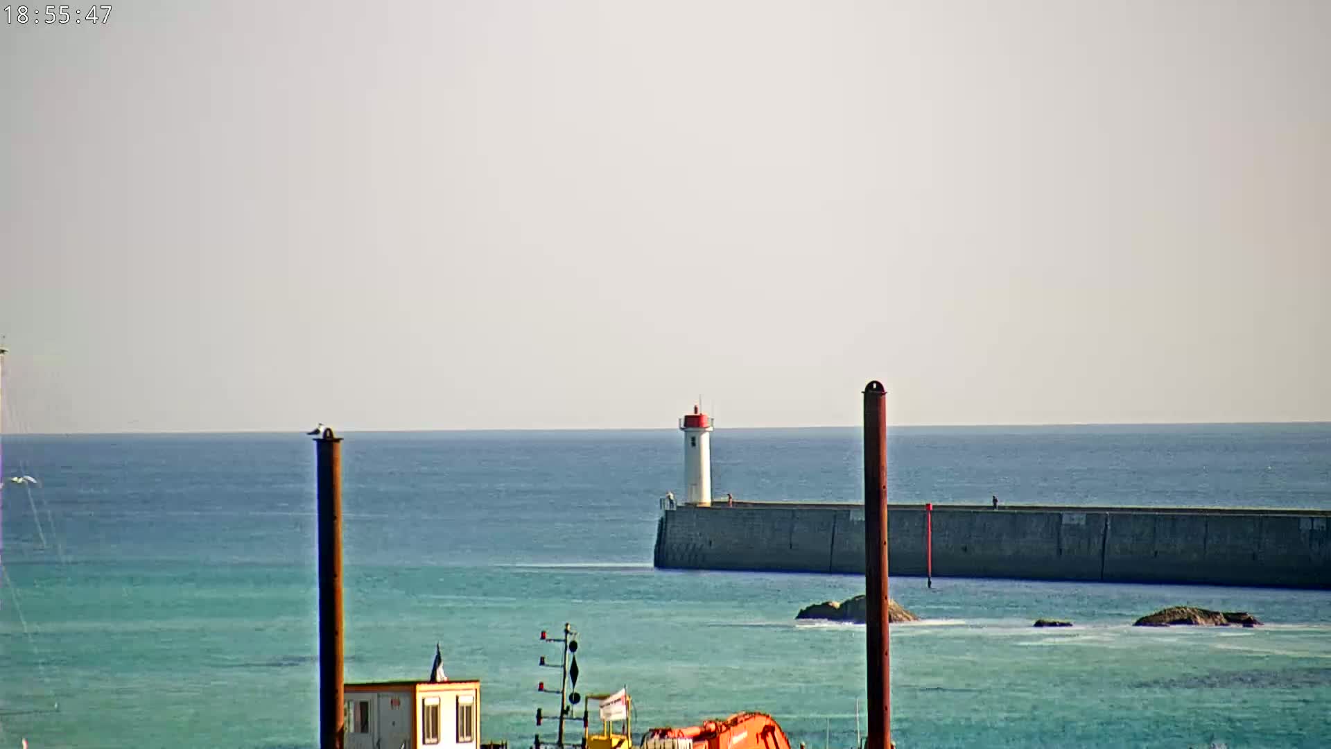 A white lighthouse stands atop a long, dark gray seawall extending into a calm, teal ocean under a bright, clear sky.