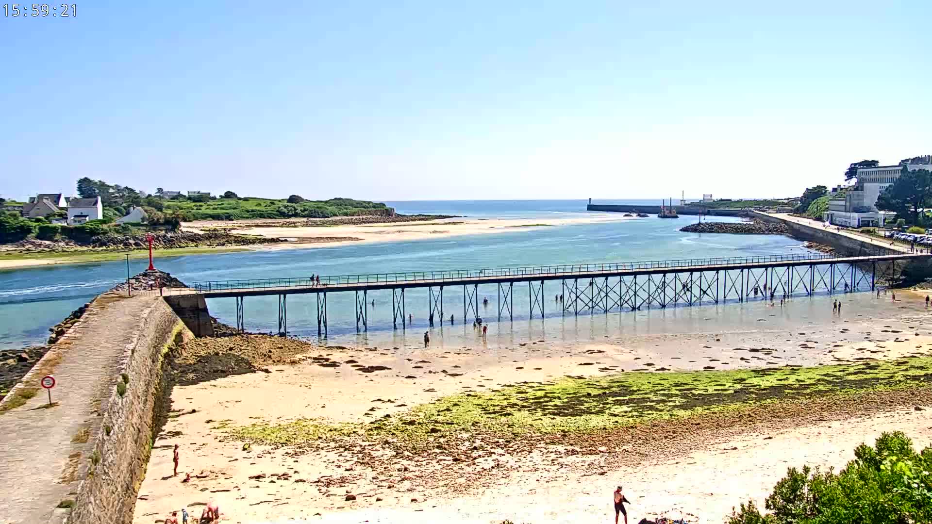 A long pier extends over shallow, clear water toward a small island on a sunny day, with people wading in the water and relaxing on the sandy shore.