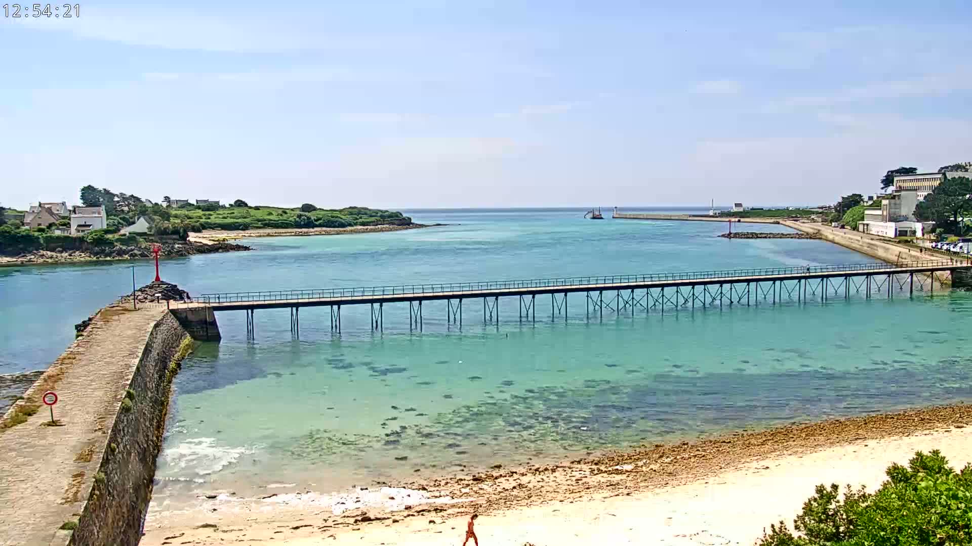 A long pier extends over clear, shallow turquoise water toward a small island under a sunny sky.