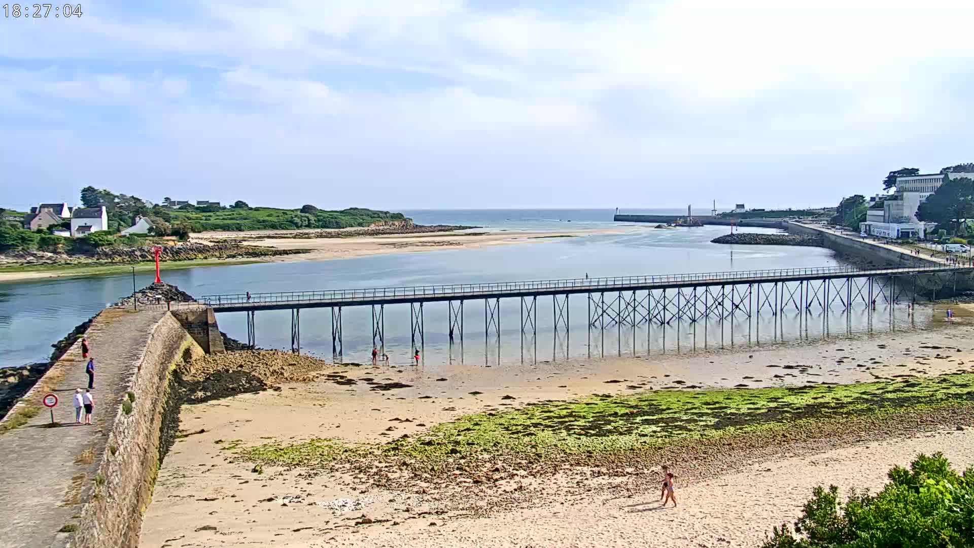 A long pier extends over calm, shallow water toward a sandy beach under a mostly sunny sky.
