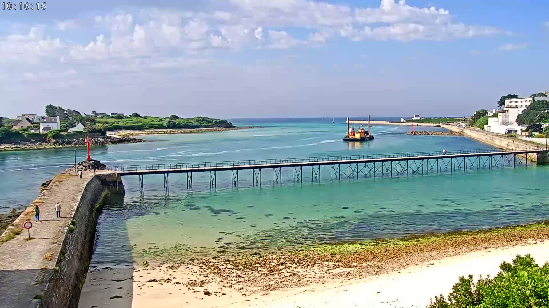 A long pier extends over clear, shallow turquoise water toward a small island under a partly cloudy sky.