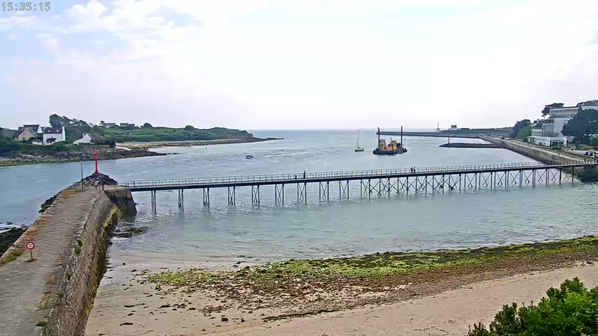 A long pier extends over calm water toward a small island and a larger landmass under a mostly cloudy sky.