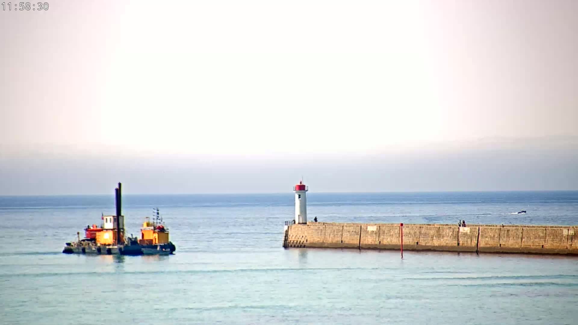 A small work boat is approaching a breakwater with a lighthouse on a calm, light-blue sea under a hazy sky.