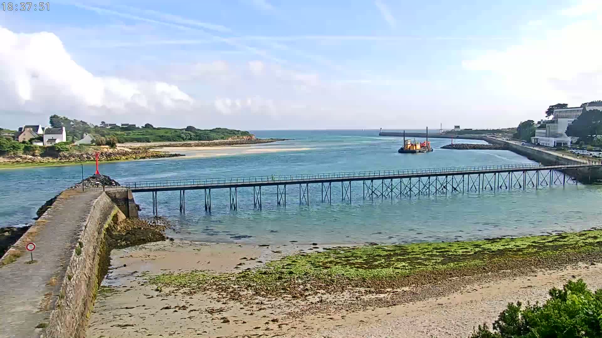 A long pier extends over calm, shallow turquoise water toward a small island under a partly cloudy sky.