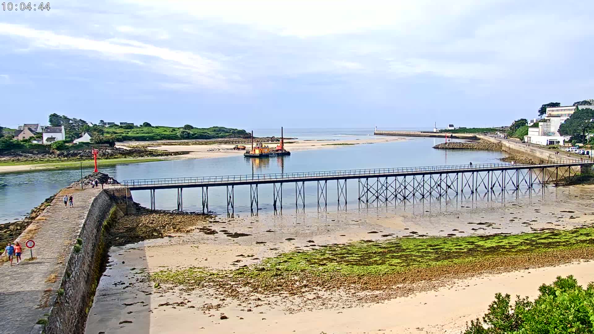 A long pier extends over a calm, shallow waterway with a sandy beach and patches of green algae, under a partly cloudy sky.